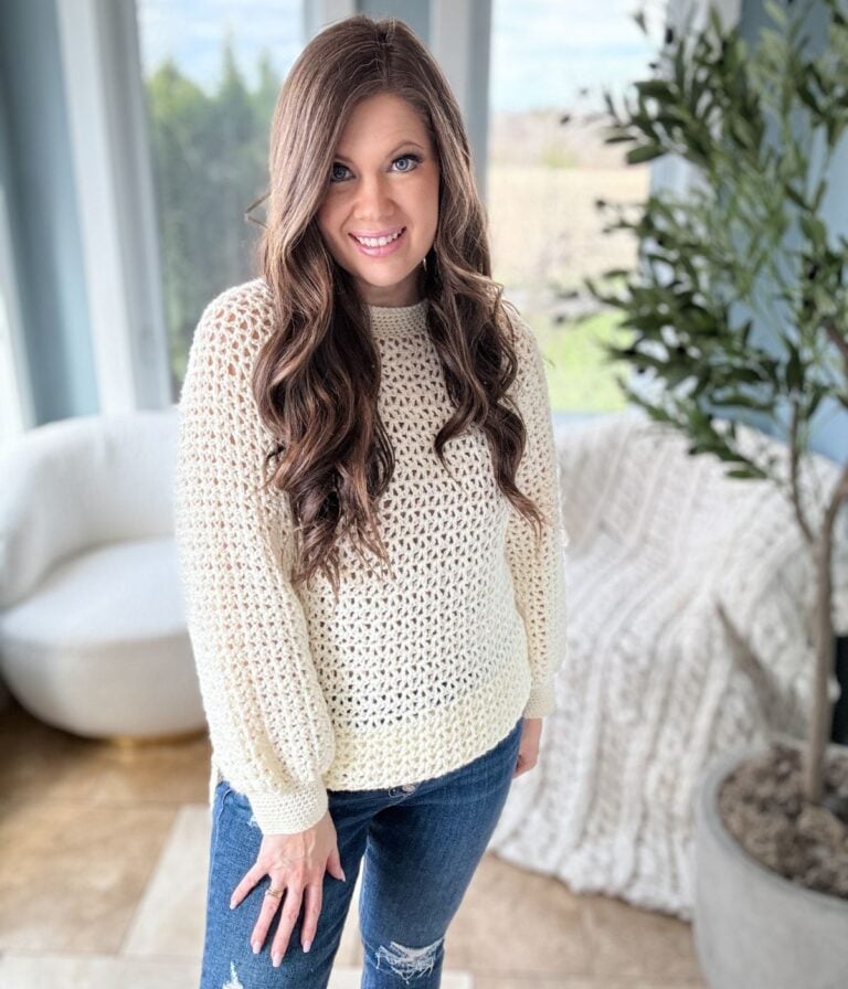 A woman with long, wavy hair wearing a cream-colored 1 Piece V Stitch Pullover and distressed jeans smiles in a bright, plant-decorated room.