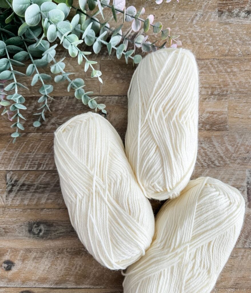 Three skeins of cream-colored yarn next to a spray of artificial greenery on a wooden surface, ideal for creating a 1 Piece V Stitch Pullover.