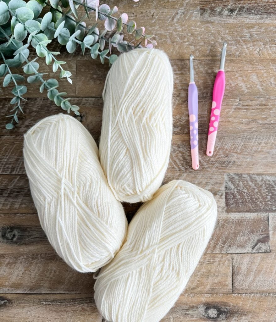 Two skeins of cream-colored yarn with pink and white crochet hooks beside them, set on a wooden surface with green eucalyptus leaves for crafting a 1 Piece V Stitch Pullover.