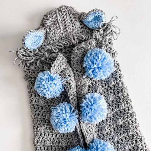 A grey and blue crocheted sleeping bag with hooded lamb blanket feature and blue pom poms.