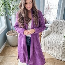 A woman with long hair stands indoors wearing a Mauve-A-Lous Duster Cardigan over a white top and jeans. A potted plant and a knitted blanket on a chair are in the background.