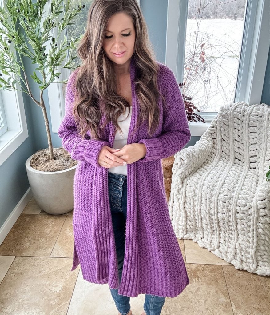A woman with long hair stands indoors wearing a Mauve-A-Lous Duster Cardigan over a white top and jeans. A potted plant and a knitted blanket on a chair are in the background.
