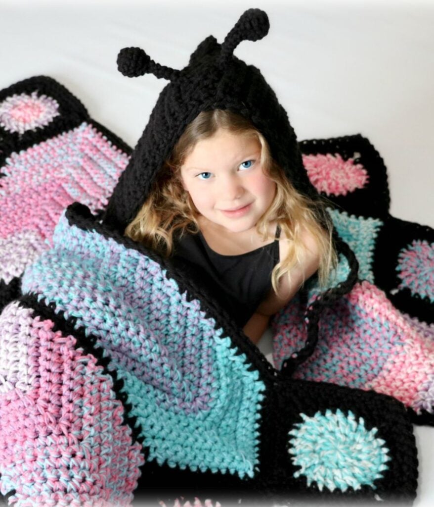 A young girl sits wrapped in a Butterfly Blanket, crocheted in pastel pink, blue, and black, with a black hood featuring antennae.