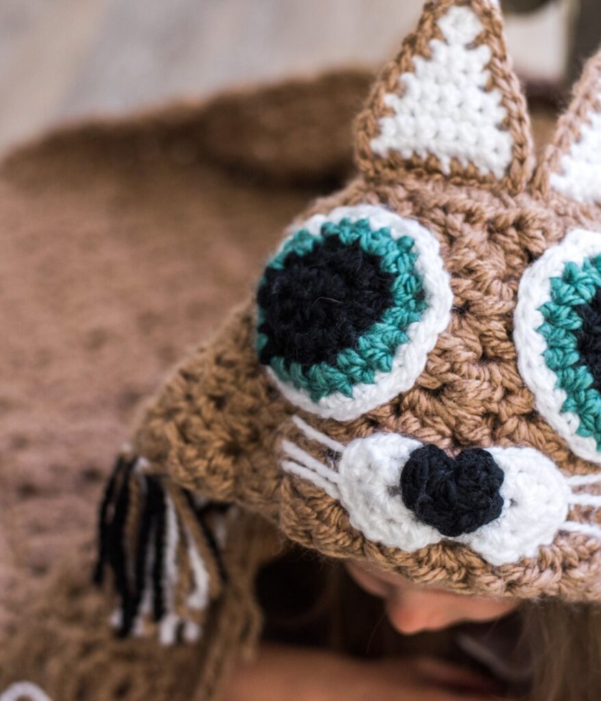 A child wearing a crocheted hat designed to look like a cat with large green and black eyes, white snout, and ears, cozily wrapped in a hooded Cat Blanket.