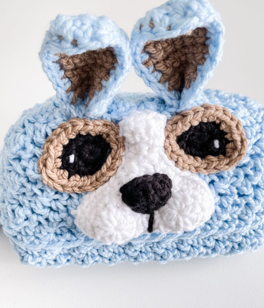 A crocheted blue and white Crochet Hooded Dog Blanket features large brown and black eyes, a black nose, and upright ears resembling a cute cartoon dog or puppy face.