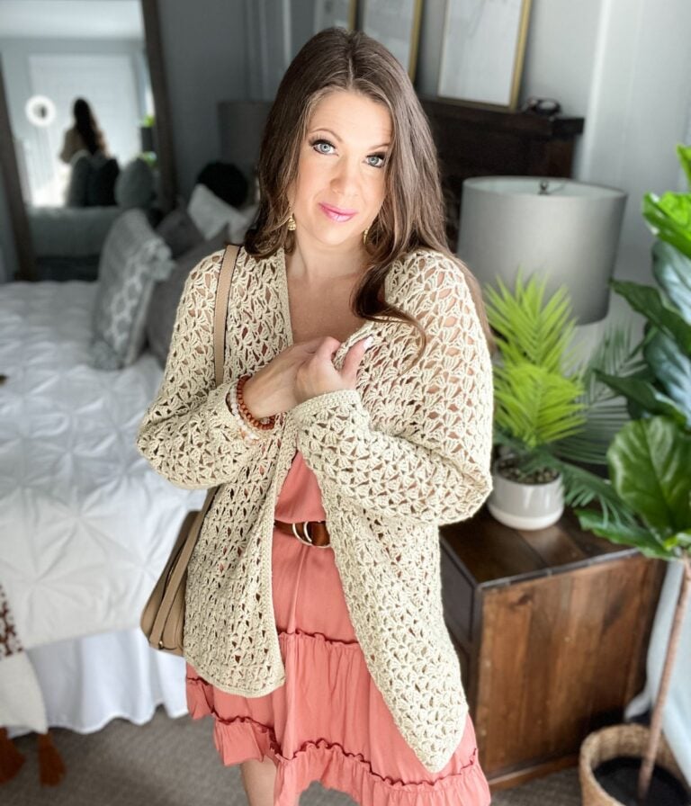 A woman in a peach dress and a beige lacy crocheted cardigan stands in a bedroom, with a reflection visible in a mirror behind her.