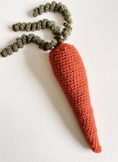 Hand-crocheted carrot with green top on a white surface, accompanied by a Crochet Hooded Rabbit Blanket.