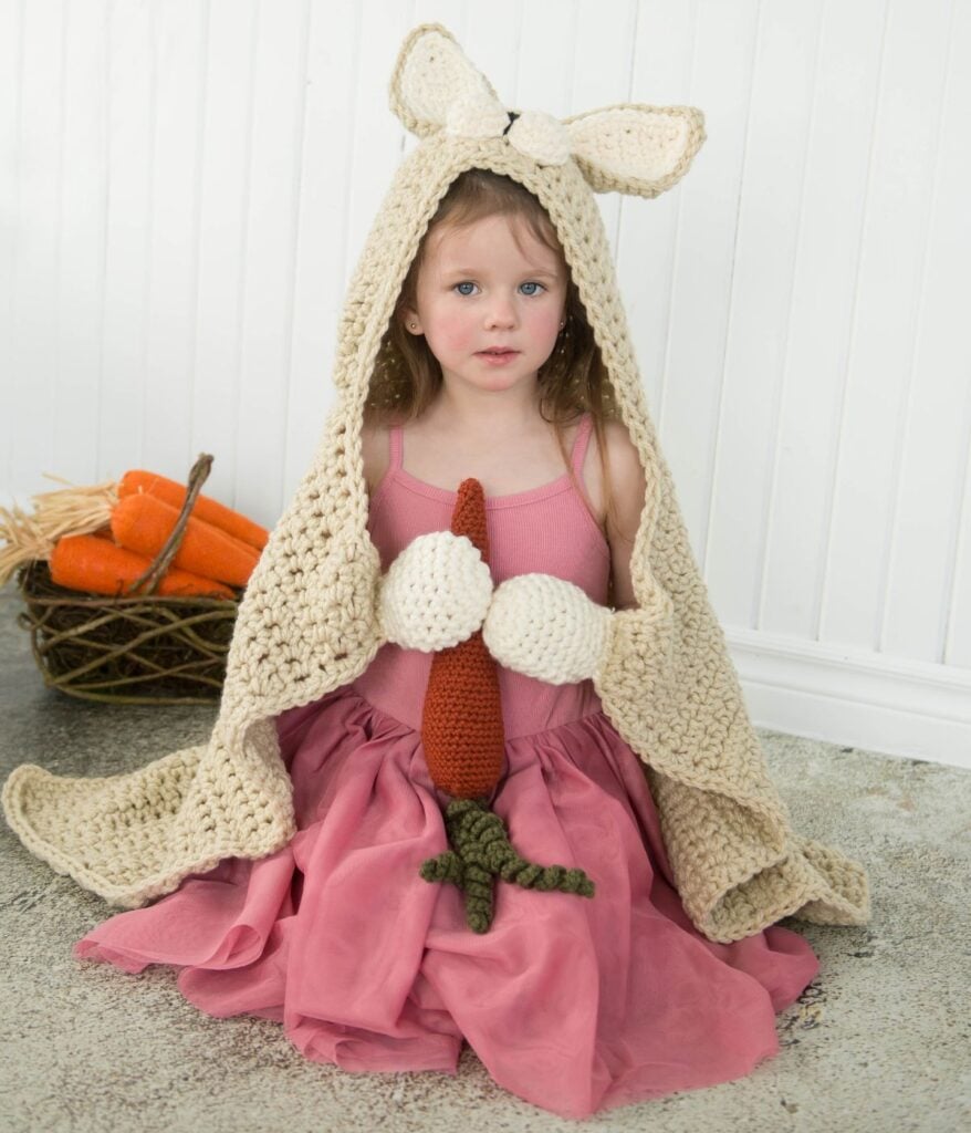 A young child dressed in a pink dress and wearing a beige crochet bunny blanket with long ears.