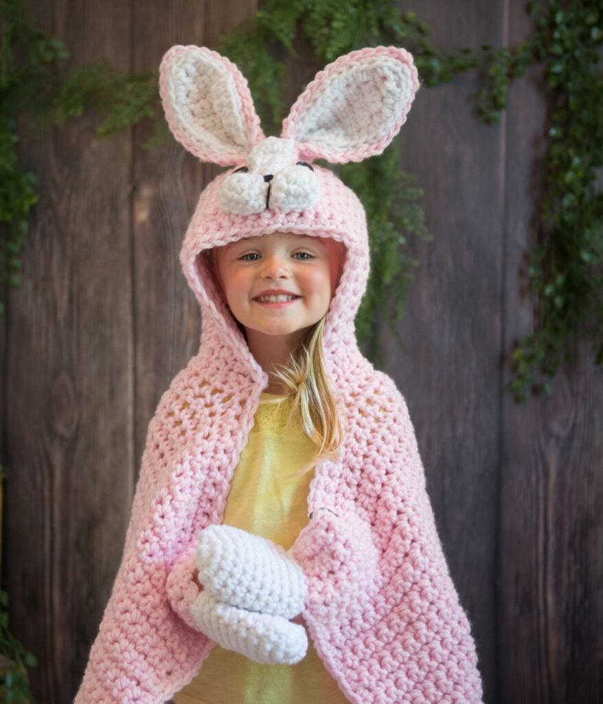 A smiling child wearing a cozy pink bunny costume with a crochet bunny blanket, featuring large ears and a hood.