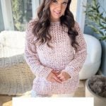 Woman wearing a pink crochet pullover stands indoors, hands clasped, with a plant and window in the background. Channel your creativity with this Gum Drop Pullover or pair it with the Lacy Days Kimono, both featuring Crochet Pattern & Video, Sizes XS-5X.