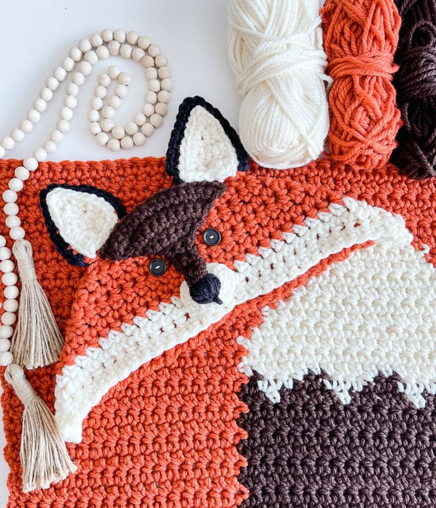 Crocheted fox design in orange, white, and brown yarn with black button eyes, beside skeins of yarn and wooden beads with a tassel—perfect for crafting your own Hooded Fox blanket.