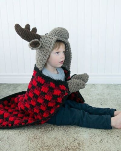A little boy wearing a crocheted reindeer hat, resembling a hooded bear blanket.