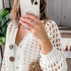 Woman taking a selfie with her smartphone, showcasing her white crochet cardigan, manicured nails, and a ring.
