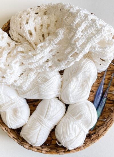 A basket containing white yarn and a crochet project to crochet a cardigan with a pair of crochet hooks.