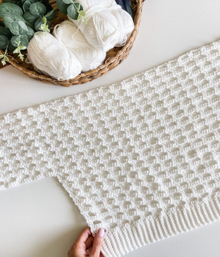 Handcrafted white crochet cardigan beside skeins of yarn and a woven basket.