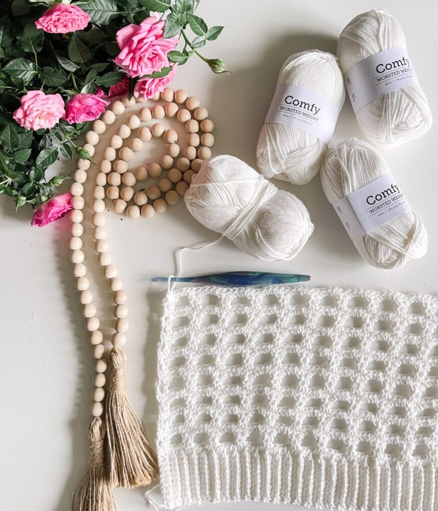 A flat lay of crochet supplies for a cardigan with white yarn, a crochet piece, a hook, and decorative wooden beads next to pink roses.