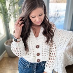 Woman in a white crochet cardigan and blue jeans standing indoors with a snowy landscape visible through the window.