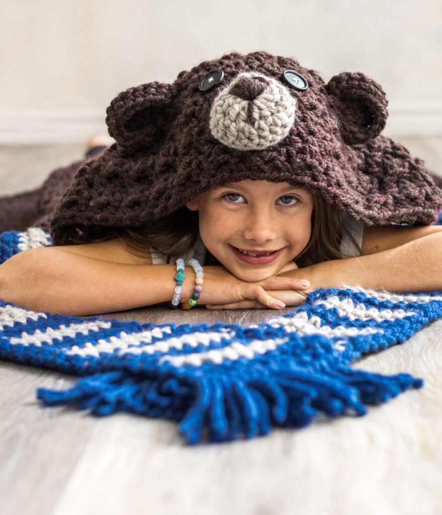 A child lies on the floor smiling, wearing a crocheted bear hood and resting on a cozy Hooded Bear Blanket with blue and white stripes.