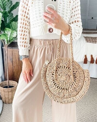 A woman wearing a beige sweater and wide leg pants carries a crochet bag.