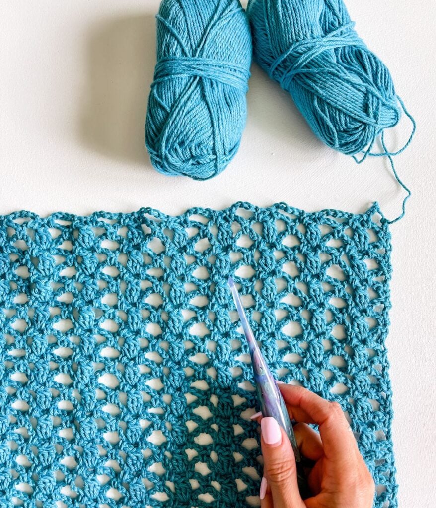 A person's hand with pastel nail polish holding crochet hooks, working on a light blue Crochet Cabot Trail Cardigan project with yarn skeins nearby.