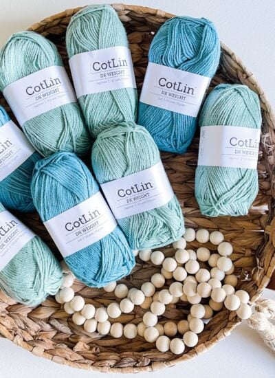 Skeins of cotlin dk weight yarn in varying shades of blue arranged in a wicker basket with a string of wooden beads for crocheting a Cabot Trail Cardigan.