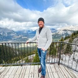 A man wearing a Mountain Ridge Cardigan stands on a wooden deck overlooking mountains.