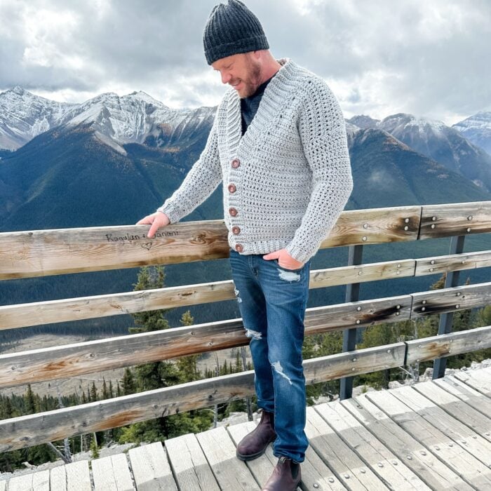 A man in a Mountain Ridge Cardigan is standing on a railing, admiring the mountains.