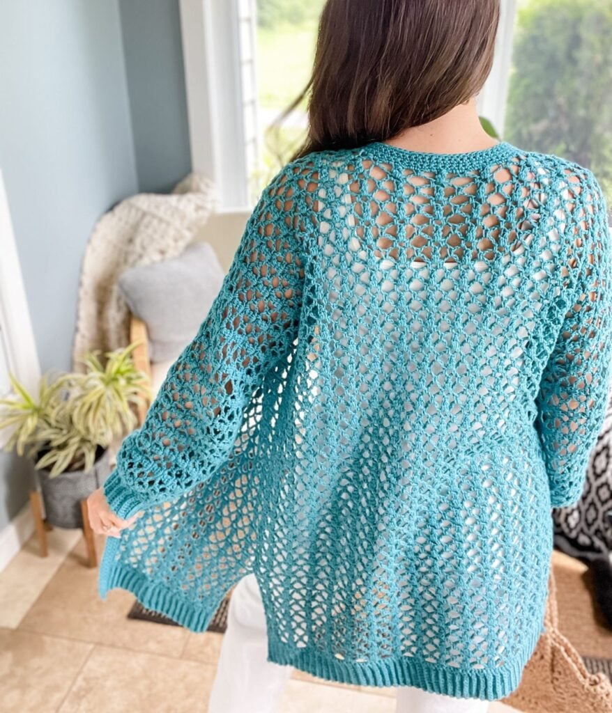 Woman wearing a teal Crochet Cabot Trail Cardigan with an openwork pattern standing in a room with plants and a chair.