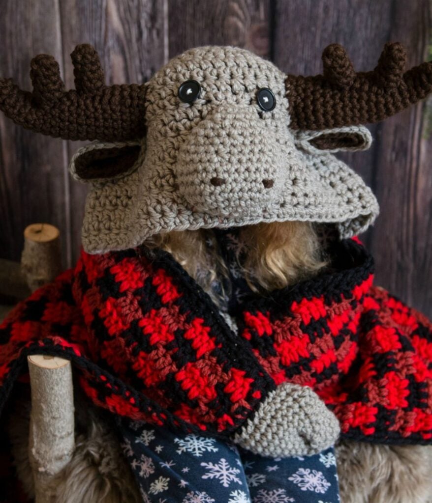 Person wearing a crocheted moose hat, wrapped in a red and black plaid Crocheted Hooded Moose Blanket, sitting on a rustic wooden chair with a wood panel background.