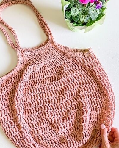A hand holding a pink crocheted tote bag with social media links.