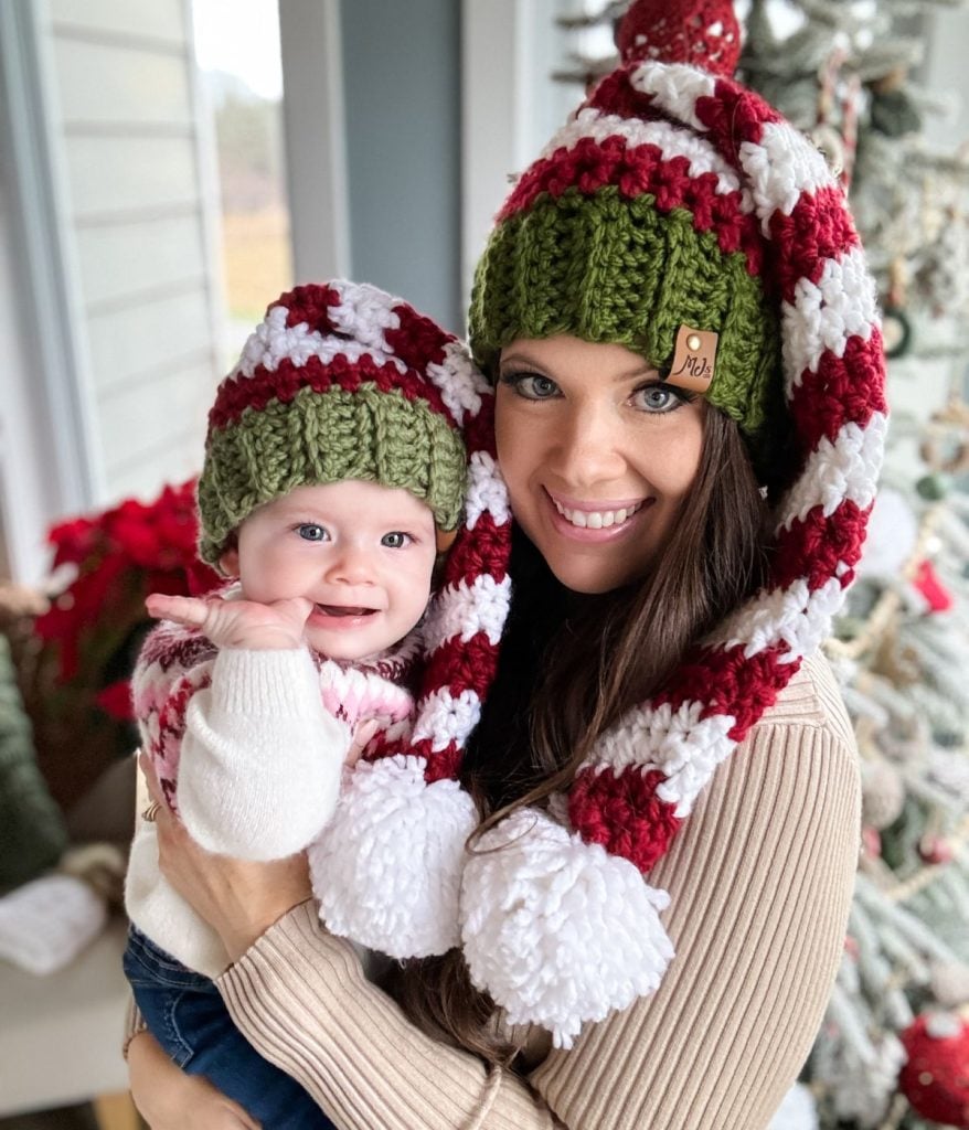 A woman and a baby, snuggled in matching red, white, and green knitted hats with pom-poms and cozy crossover wrap cardigans, beam joyful smiles indoors.