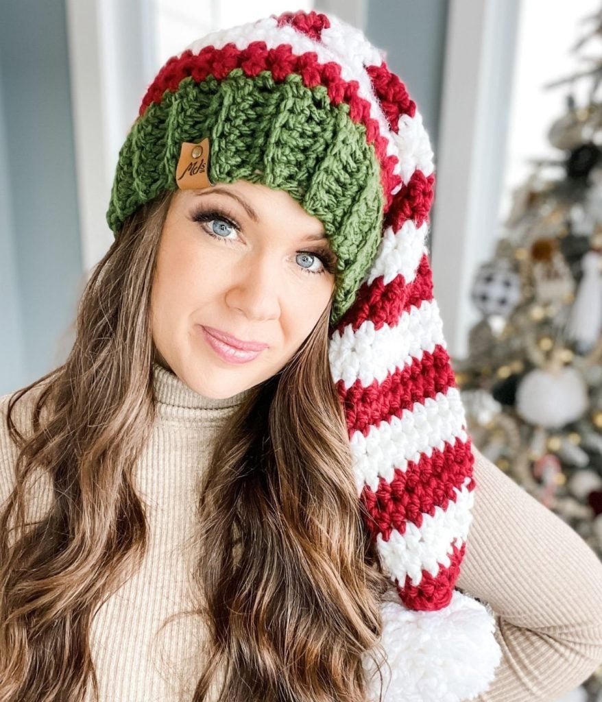 A woman wearing a striped red, white, and green knitted hat and a cozy crossover wrap cardigan poses in front of a decorated Christmas tree.