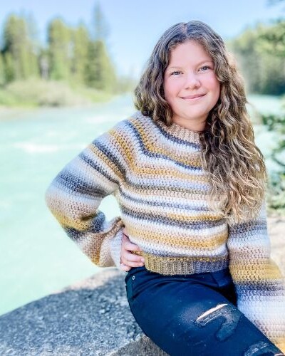 A girl sitting on a rock next to a river while wearing an easy crochet raglan sweater.