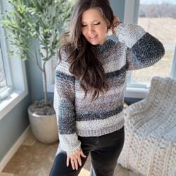 A woman in a striped, easy crochet raglan sweater smiling while standing indoors by a window.