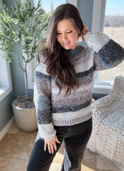 A woman in a striped, easy crochet raglan sweater smiling while standing indoors by a window.