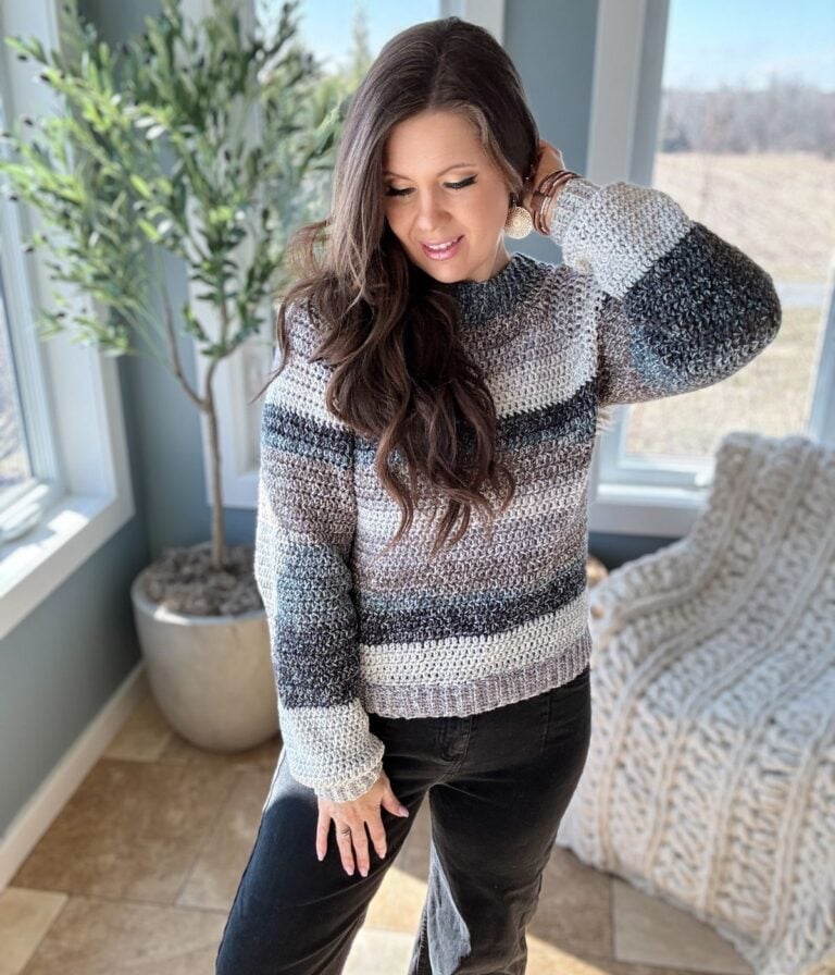 A woman in a striped, easy crochet raglan sweater smiling while standing indoors by a window.