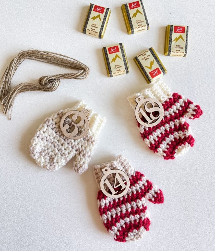 Three crocheted mittens, including a charming Mini Striped Mitten, each with a numbered wooden tag (13, 14, 18), yarn, and five small boxes of matches arranged on a white background.