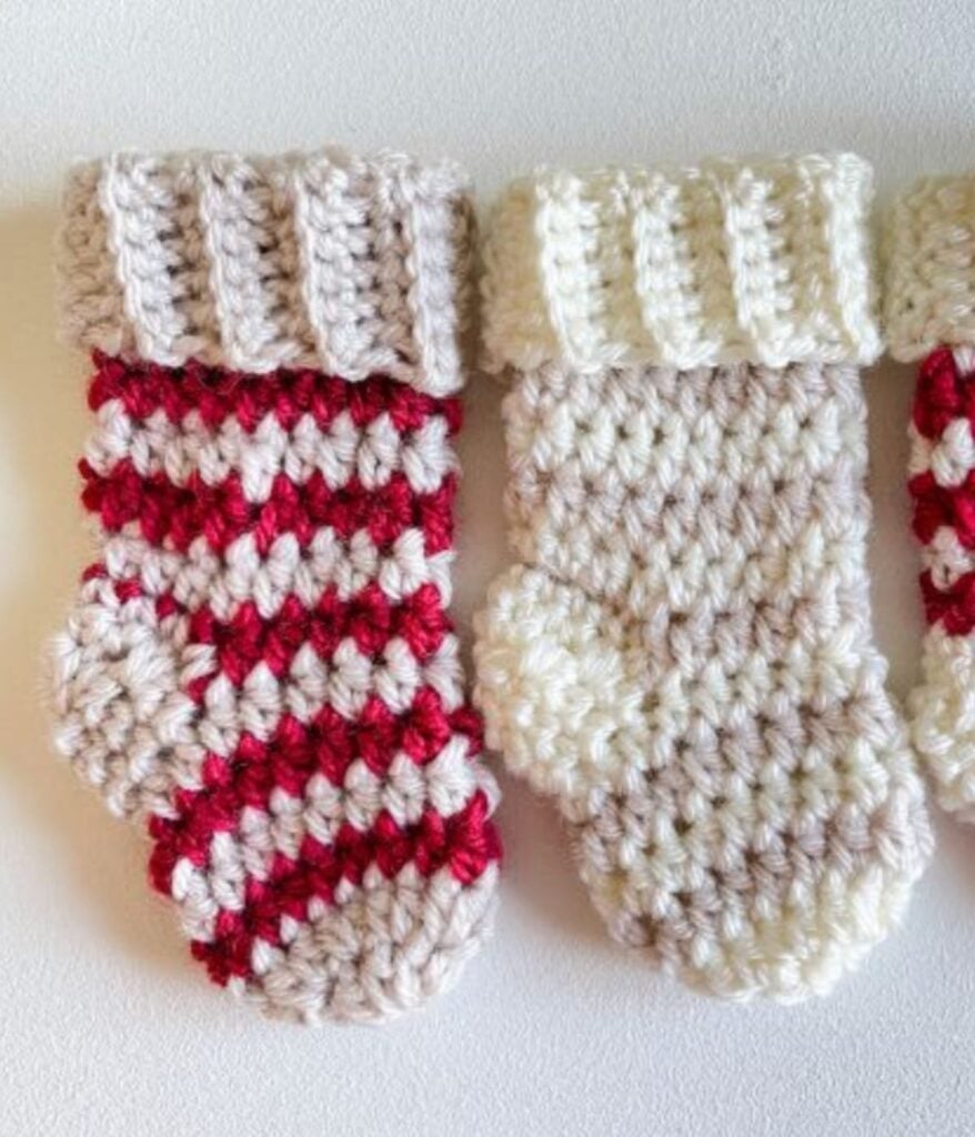 Two crocheted mini Christmas stockings, including a charming mini striped stocking in red and white and a solid off-white with a cream cuff, are placed side by side on a white surface.
