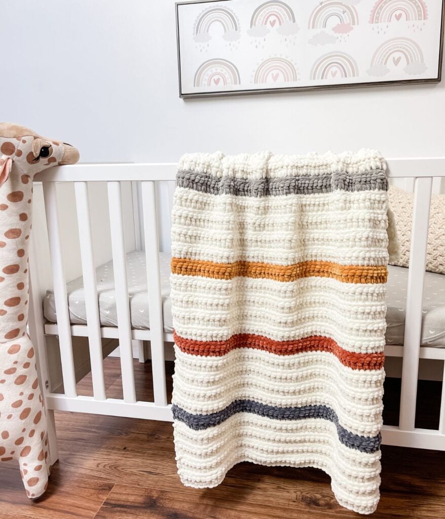 A white crib with a plush giraffe toy beside it and a ribbed baby blanket draped over the rail; rainbow artwork hangs on the wall above.