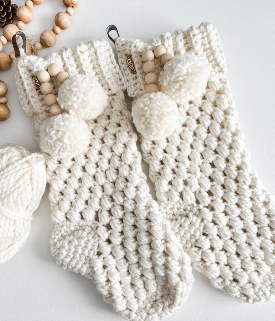 Two chunky knit white Christmas stockings, crafted in the Bulky & Quick Puff Stitch Stockings style, with pom-poms and bead decorations, lie on a white surface next to a ball of yarn and a beaded garland.