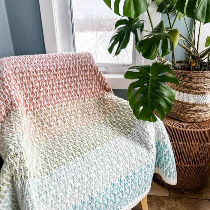 A beautiful mosaic crochet blanket sitting on a chair next to a potted plant.