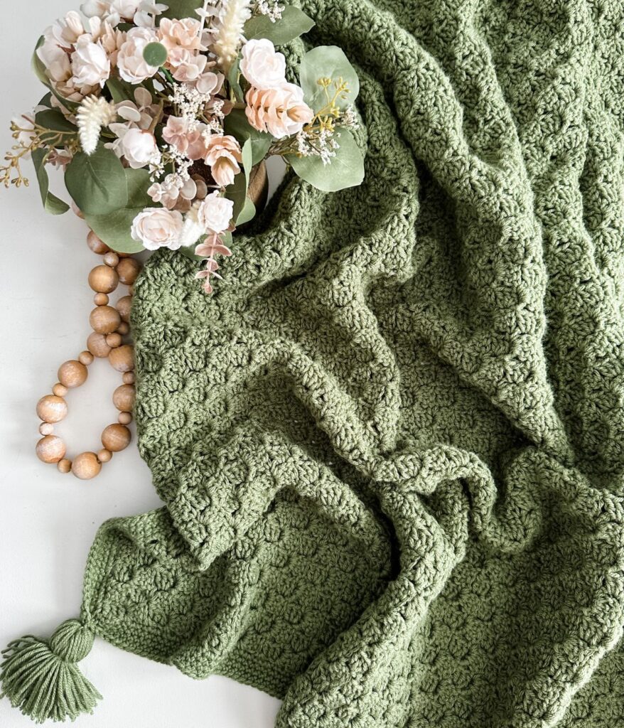 A textured green easy crochet C2C blanket with a tassel lies next to a wooden bead garland and a bouquet of pale pink and white flowers with green leaves.