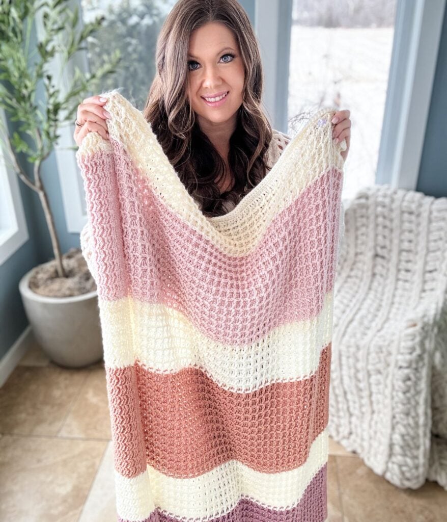 A woman stands indoors holding up a striped Waffle Stitch Blanket in shades of pink, cream, and brown. A potted plant and another blanket are visible in the background.