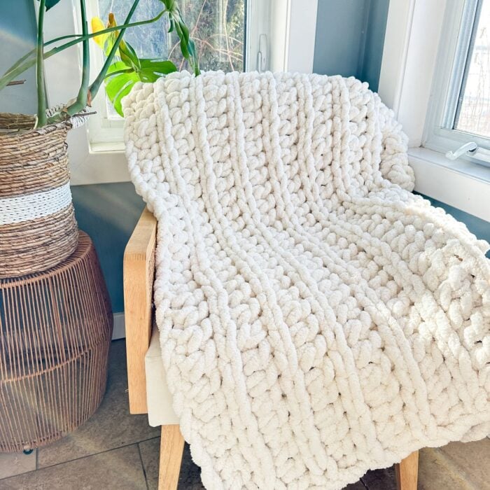 A chunky white knitted blanket sitting on a chair in front of a window.