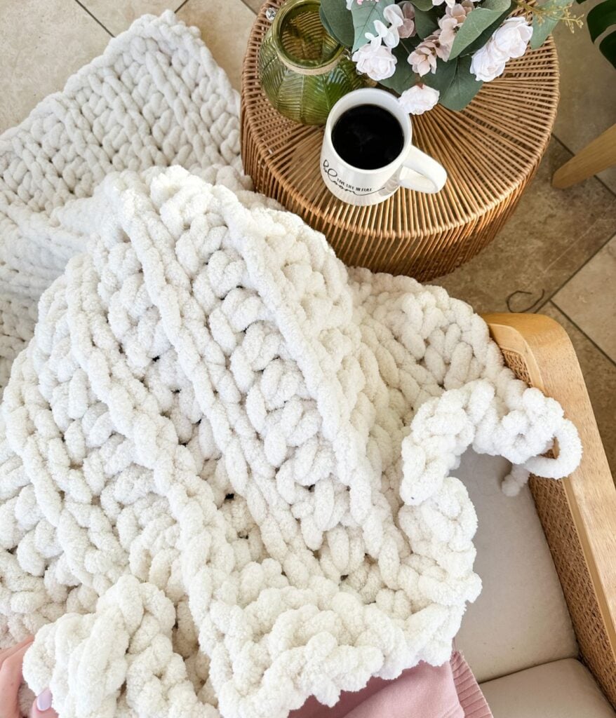 A chunky throw knit blanket draped over a chair next to a wicker table with a cup of black coffee, a green vase, and a pink flower arrangement.
