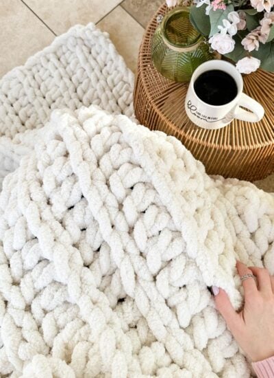 A person is holding a chunky white knit throw on a rug.