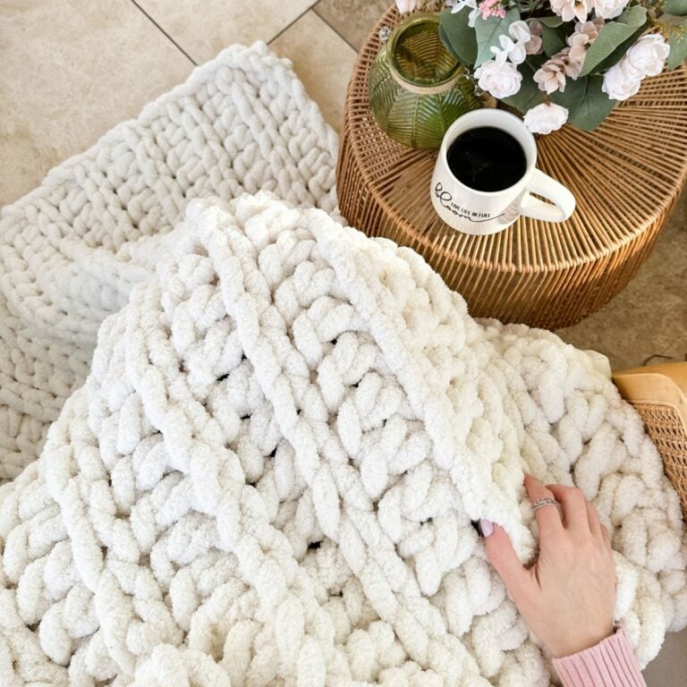 A person is holding a chunky white knit throw on a rug.