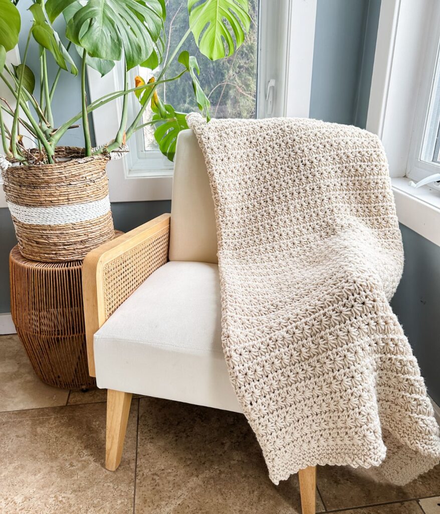 A cream-colored Star Stitch crochet blanket draped over a white chair next to a woven basket planter with a large green plant, situated by a window.