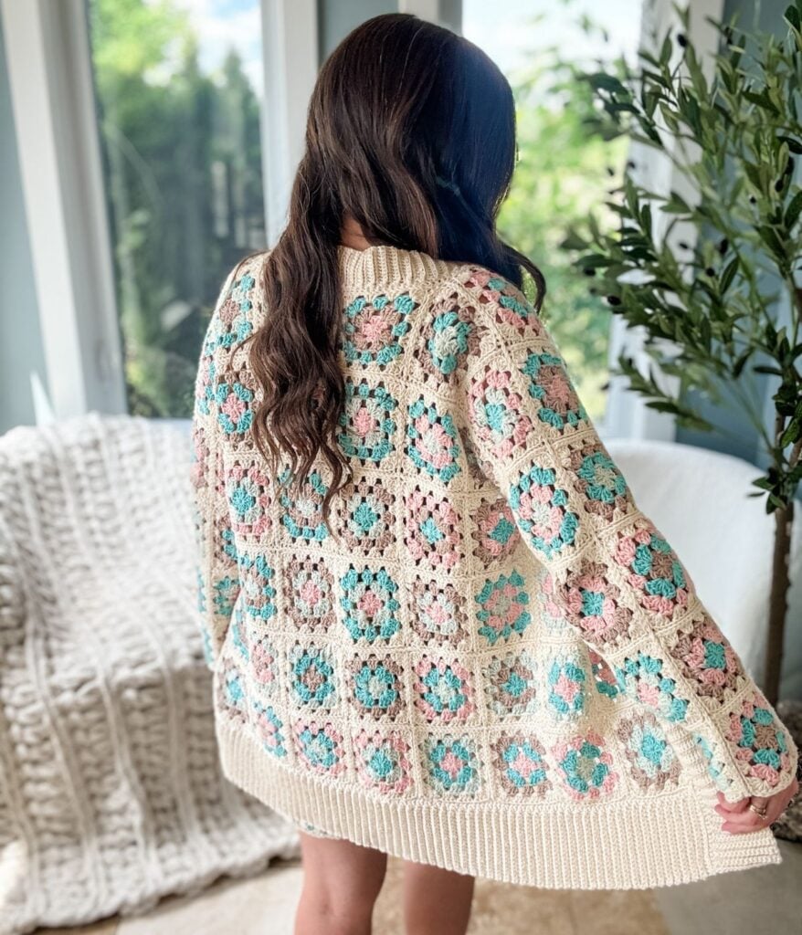 Person with long hair wearing a Granny Square Cardigan in a floral pattern, standing indoors with greenery and white knit blankets in the background.