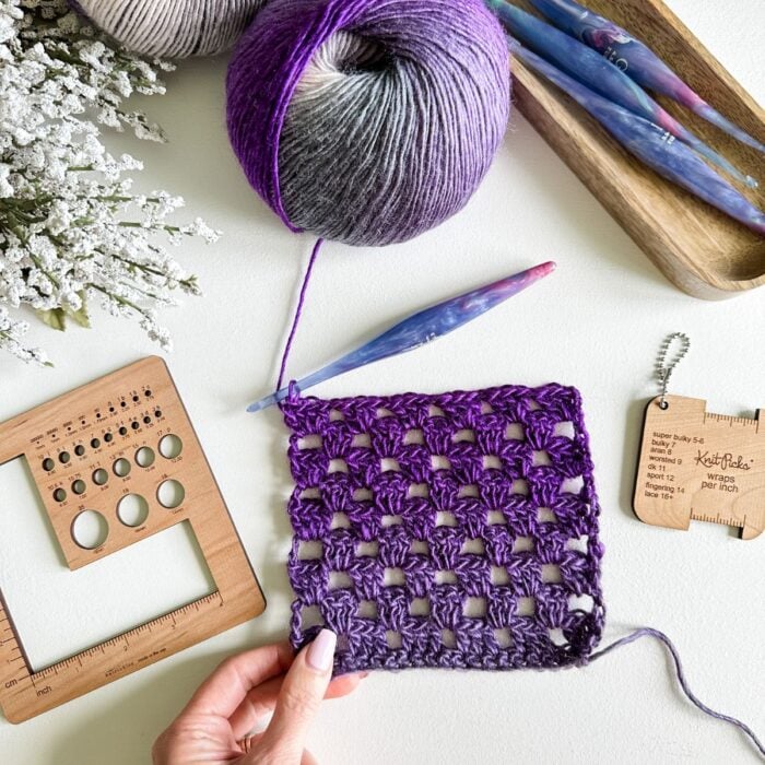 A person is crocheting a purple stitch while wearing a hooded crochet cardigan.